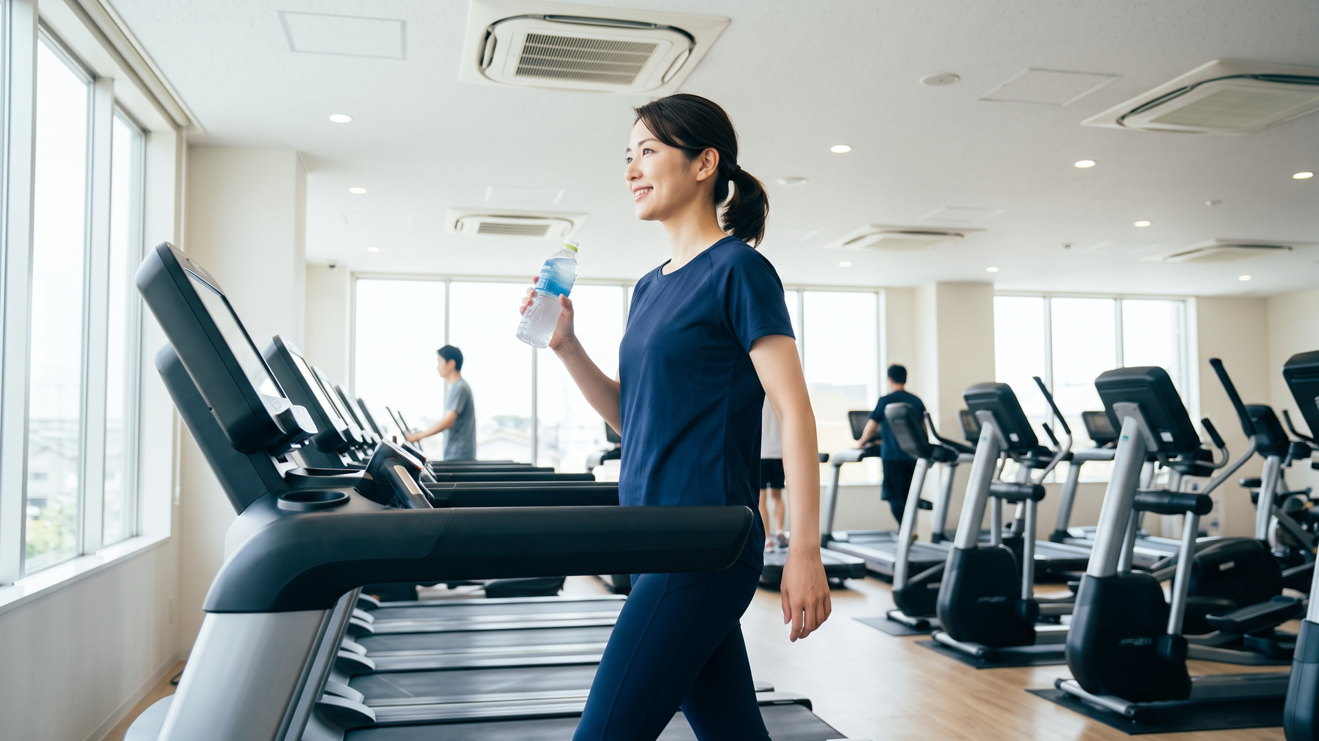 空調の効いた室内ジムでスポーツドリンクを飲みながら軽いウォーキングをする日本人女性の実写写真